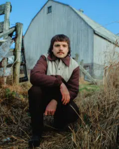 A person with a mustache squats in front of a weathered wooden barn and fence, wearing a brown and beige jacket, surrounded by dry grass under a clear blue sky.
